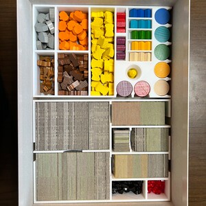 May include: A white plastic organizer box with compartments for storing game pieces. The box contains various colored wooden pieces, including gray, orange, yellow, red, blue, green, brown, and black. There are also stacks of cards in the box.