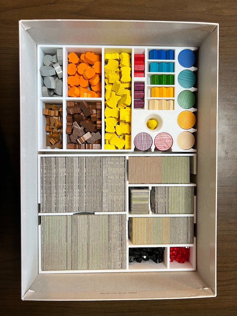 May include: A white plastic organizer box with compartments for storing game pieces. The box contains various colored wooden pieces, including gray, orange, yellow, red, blue, green, brown, and black. There are also stacks of cards in the box.