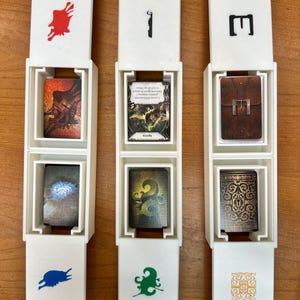 May include: Three white card holders with open lids, each containing two cards. The holders have red, black, and green symbols on the front. The cards feature fantasy art and text, with brown, green, and gold tones.