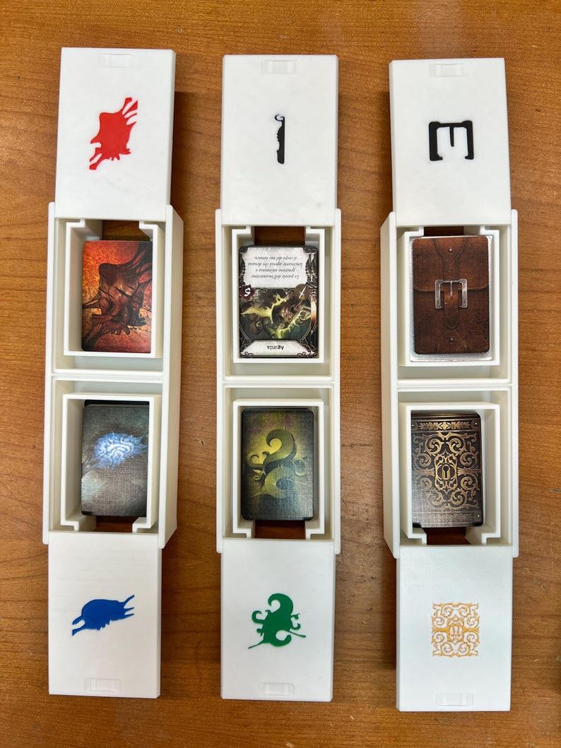 May include: Three white card holders with open lids, each containing two cards. The holders have red, black, and green symbols on the front. The cards feature fantasy art and text, with brown, green, and gold tones.