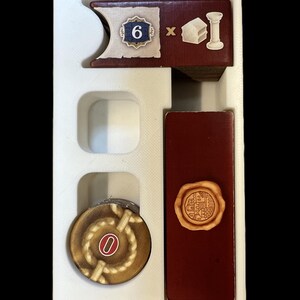 May include: A white plastic tray with a red and white card with a number 6 and a building icon, a red card with a wax seal, and a brown token with a rope design and a number 0.