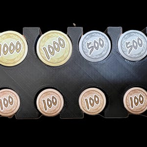 Op de afbeelding: Een zwarte plastic muntendoos met acht vakjes. De vakjes bevatten zes goudkleurige munten met de nummers 1000 en 100, en twee zilverkleurige munten met het nummer 500.
