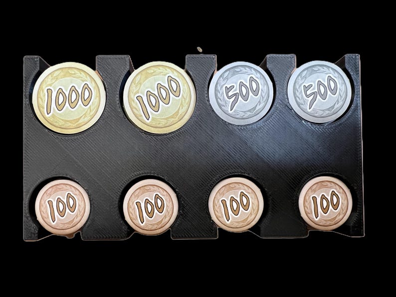 Op de afbeelding: Een zwarte plastic muntendoos met acht vakjes. De vakjes bevatten zes goudkleurige munten met de nummers 1000 en 100, en twee zilverkleurige munten met het nummer 500.
