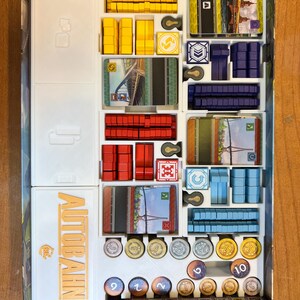 Pode incluir: Um inserto organizador branco para o jogo de tabuleiro "Autobahn" com compartimentos para as pe&ccedil;as do jogo, os cart&otilde;es e as fichas. O inserto tem uma etiqueta que diz "Autobahn" em letras douradas.