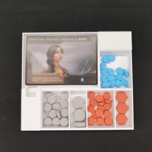 May include: A white game organiser with several compartments. It holds a card with a woman's image, blue teardrop-shaped tokens, grey circular tokens, and orange hexagonal tokens. The organiser is on a black surface.