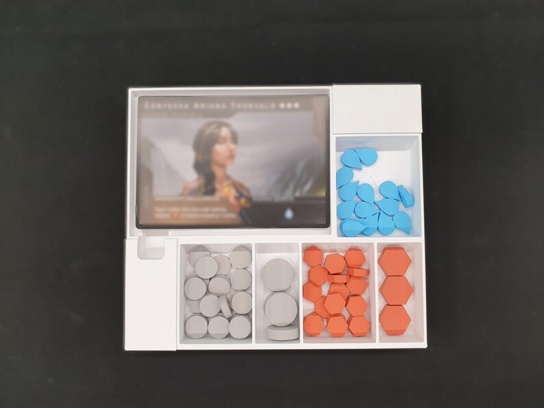 May include: A white game organiser with several compartments. It holds a card with a woman's image, blue teardrop-shaped tokens, grey circular tokens, and orange hexagonal tokens. The organiser is on a black surface.