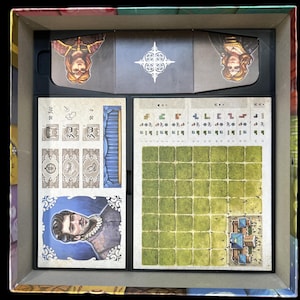 Pu&ograve; includere: Un tabellone di gioco con una griglia di quadrati, un piccolo edificio e un ritratto di un uomo con la barba. Ci sono anche diverse carte pi&ugrave; piccole con illustrazioni e testo.