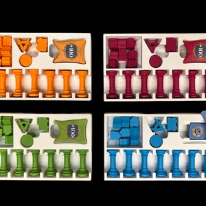 May include: Four white trays with different colored game pieces and cards. Each tray has 10 columns with small pillars in each column. The trays have a card with the text " +100" in the top right corner. The colors of the game pieces are orange, red, green, and blue.