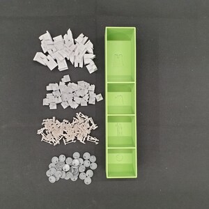 May include: A light green, four-compartment organizer with various game pieces. The pieces include white building blocks, tan chairs, and gray circular tokens. The organizer has etched images in each compartment.