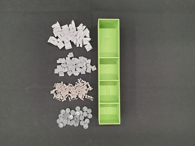 May include: A light green, four-compartment organizer with various game pieces. The pieces include white building blocks, tan chairs, and gray circular tokens. The organizer has etched images in each compartment.