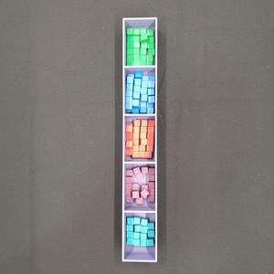 May include: A white rectangular container divided into five sections, each filled with small, colourful cubes. The cubes are green, blue, orange, pink, and teal. The container is on a dark grey surface.