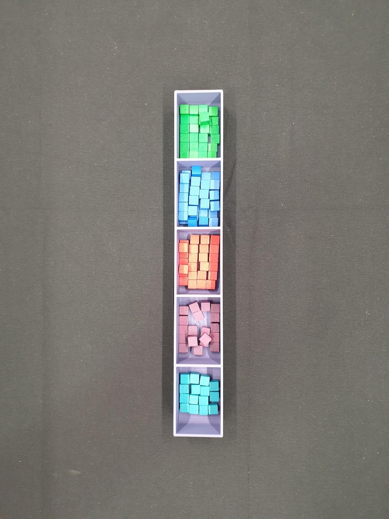 May include: A white rectangular container divided into five sections, each filled with small, colourful cubes. The cubes are green, blue, orange, pink, and teal. The container is on a dark grey surface.