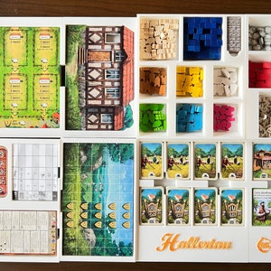 May include: A white organizer with compartments for storing game pieces and cards. The organizer has a white background with a brown and white house illustration. The organizer is labeled "Hallertau".