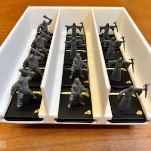 May include: A white plastic organizer filled with miniature gray figurines. The figurines are of various characters, including warriors with weapons and robed figures. The organizer sits on a wooden surface.