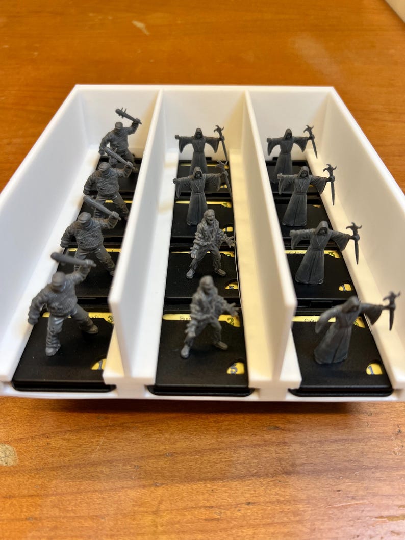 May include: A white plastic organizer filled with miniature gray figurines. The figurines are of various characters, including warriors with weapons and robed figures. The organizer sits on a wooden surface.