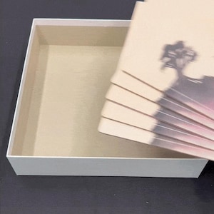 以下が含まれることがあります： ベージュの内装の白い長方形の箱。風景デザインのベージュ色のカードの束が箱の隣に部分的に見えています。カードには木と山のシルエットがあります。