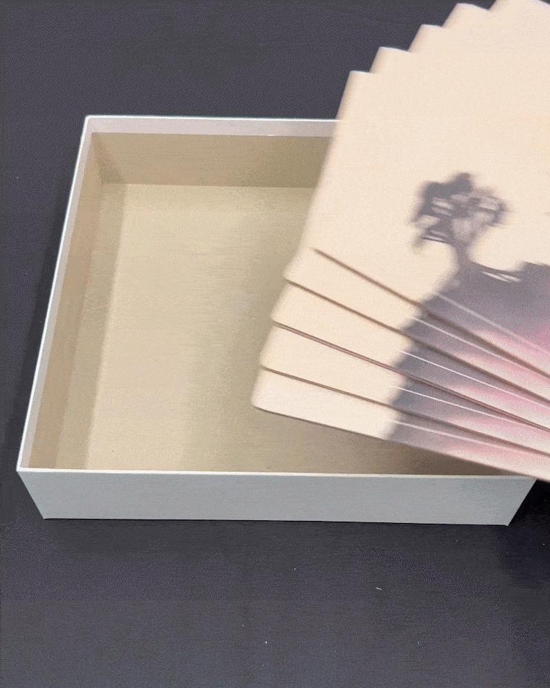 以下が含まれることがあります： ベージュの内装の白い長方形の箱。風景デザインのベージュ色のカードの束が箱の隣に部分的に見えています。カードには木と山のシルエットがあります。