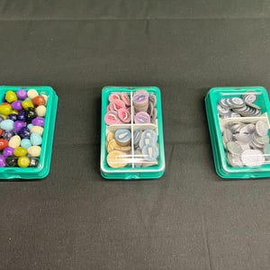 以下が含まれることがあります： ゲームの駒が入った3つの緑色のプラスチック容器。1つの容器には、カラフルな丸いゲームの駒が入っています。2番目の容器には、ピンク、青、タン色のゲームの駒が入っています。3番目の容器には、銀色のゲームの駒が入っています。