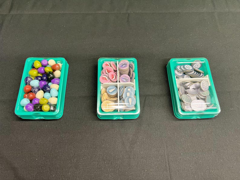 以下が含まれることがあります： ゲームの駒が入った3つの緑色のプラスチック容器。1つの容器には、カラフルな丸いゲームの駒が入っています。2番目の容器には、ピンク、青、タン色のゲームの駒が入っています。3番目の容器には、銀色のゲームの駒が入っています。