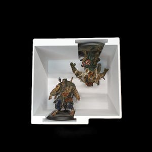 May include: Two detailed miniature figurines of fantasy characters are displayed inside a white box against a black background. One figure stands upright, while the other is positioned upside down. The figures are painted with various colors and appear to be part of a tabletop game.