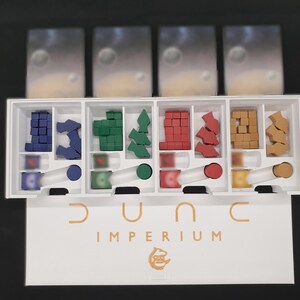 May include: A board game organiser for Dune: Imperium. The white box is divided into compartments holding various game pieces in blue, green, red, and brown. The game title "Dune Imperium" is printed in gold on the box.