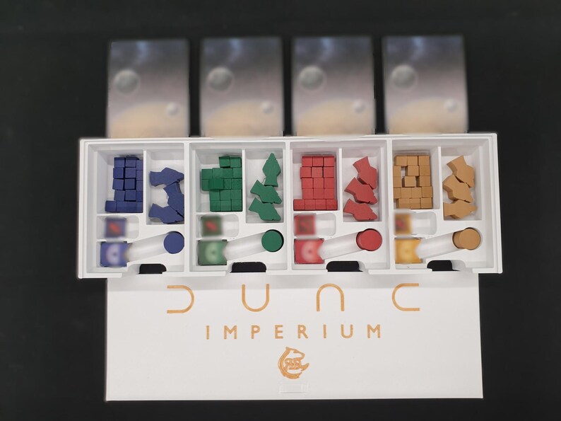 May include: A board game organiser for Dune: Imperium. The white box is divided into compartments holding various game pieces in blue, green, red, and brown. The game title "Dune Imperium" is printed in gold on the box.