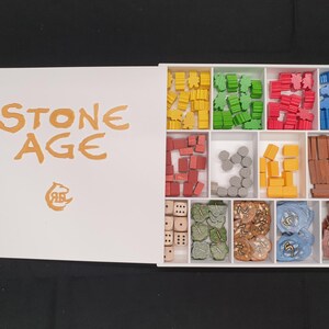 Puede incluir: Caja blanca abierta del juego de mesa "Stone Age" con piezas de juego de colores. La caja contiene compartimentos con piezas amarillas, verdes, rojas y azules, adem&aacute;s de dados y otros componentes del juego.