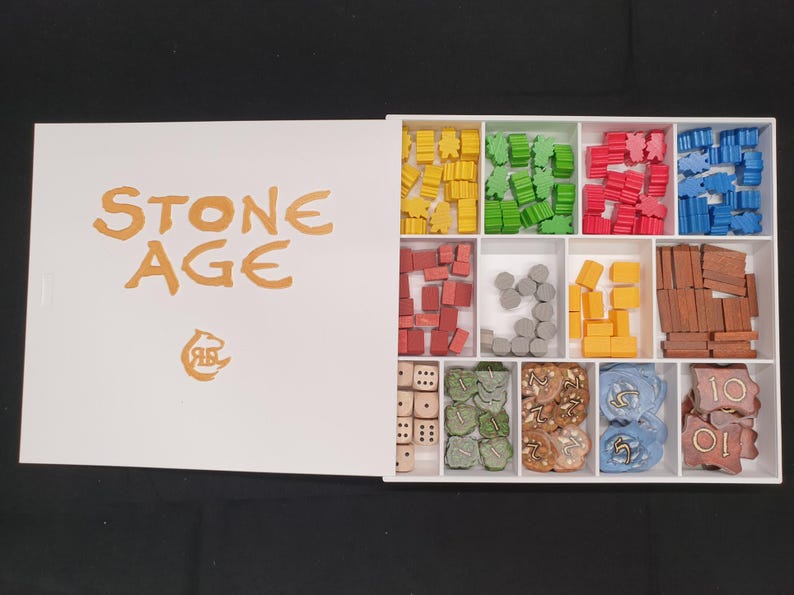 Puede incluir: Caja blanca abierta del juego de mesa "Stone Age" con piezas de juego de colores. La caja contiene compartimentos con piezas amarillas, verdes, rojas y azules, adem&aacute;s de dados y otros componentes del juego.