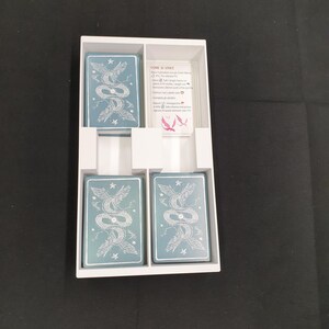 May include: A white card game box containing four decks of cards and a rules card. The cards feature a teal background with a white illustration of two intertwined serpents. The rules card has Italian text and a pink illustration.