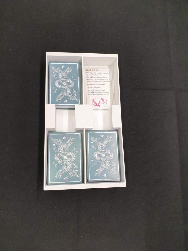 May include: A white card game box containing four decks of cards and a rules card. The cards feature a teal background with a white illustration of two intertwined serpents. The rules card has Italian text and a pink illustration.