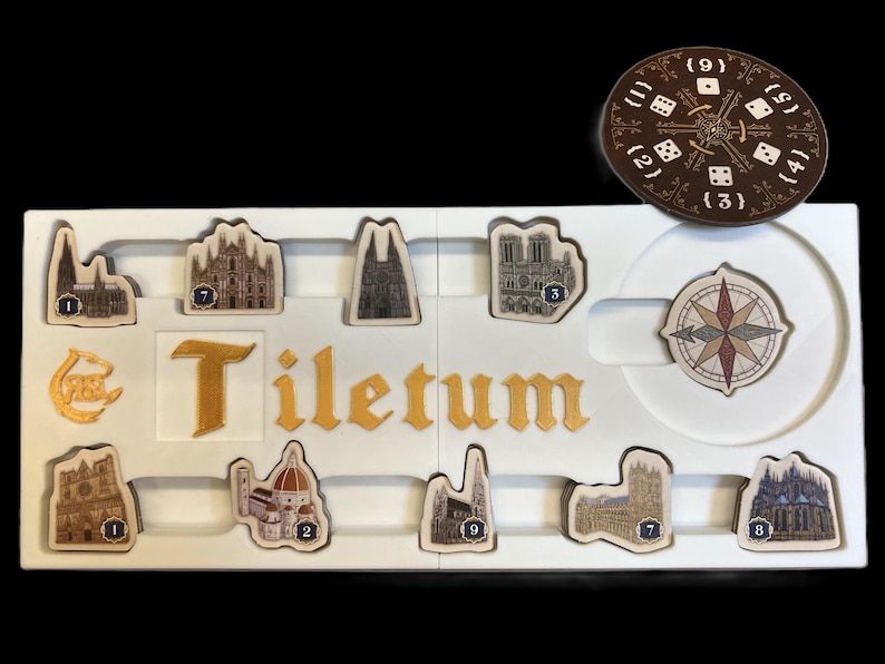 May include: A white game board with the word "Tiletum" printed in gold letters. The board has nine slots for game pieces, each with a number from 1 to 9. There are also two additional slots for a compass and a die. The game pieces are small, detailed images of buildings.