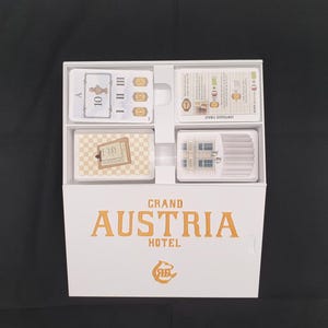 May include: A white box containing a board game titled "Grand Austria Hotel." The box is open, revealing four compartments filled with cards. The cards feature various designs, including numbers, illustrations, and text. The game's logo is in orange.