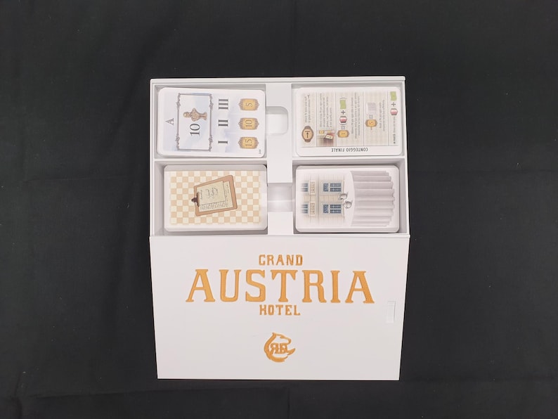 May include: A white box containing a board game titled "Grand Austria Hotel." The box is open, revealing four compartments filled with cards. The cards feature various designs, including numbers, illustrations, and text. The game's logo is in orange.