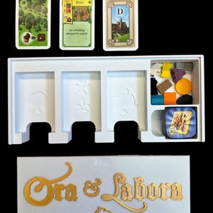 May include: A white game box with the words "Ora & Labora" in gold lettering. The box contains a white insert with three slots for cards and a compartment for game pieces. There are three cards visible: a green card with a forest, a yellow card with a building, and a brown card with a castle. The game pieces include wooden pieces in various colors and shapes.