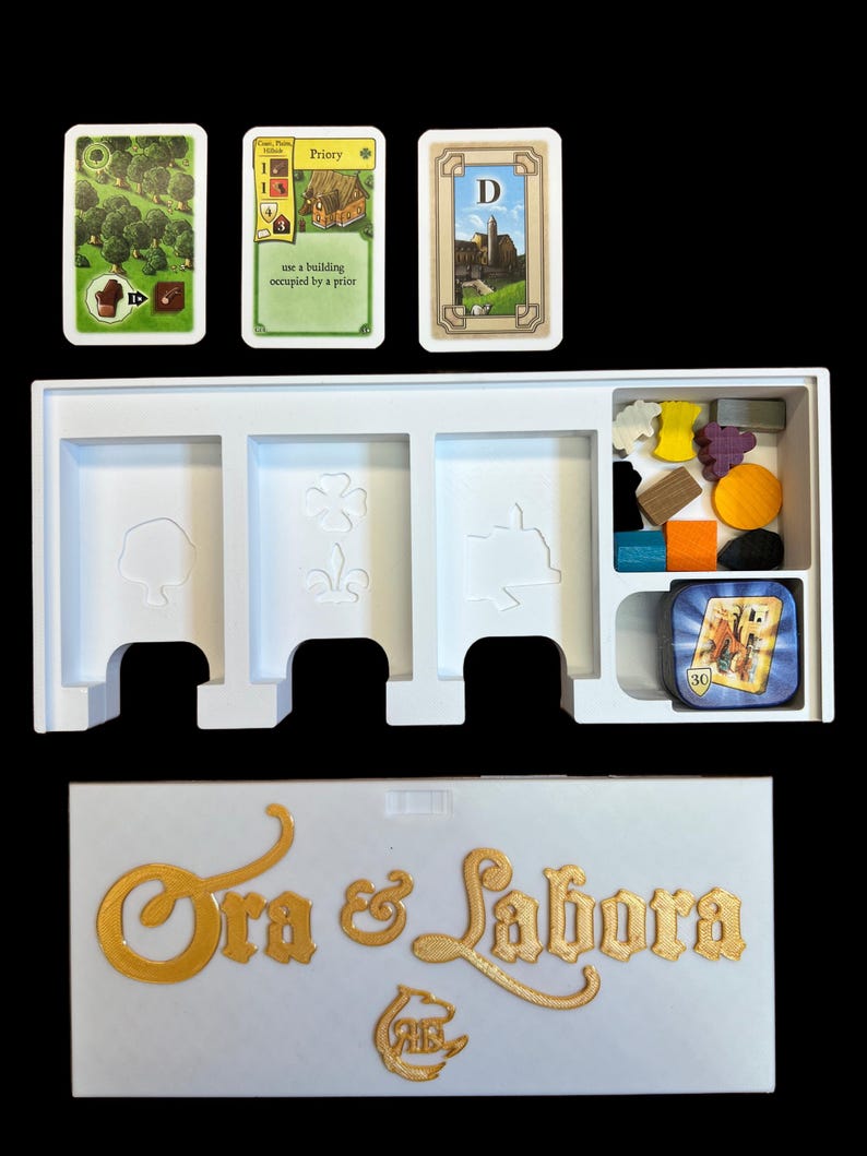 May include: A white game box with the words "Ora & Labora" in gold lettering. The box contains a white insert with three slots for cards and a compartment for game pieces. There are three cards visible: a green card with a forest, a yellow card with a building, and a brown card with a castle. The game pieces include wooden pieces in various colors and shapes.