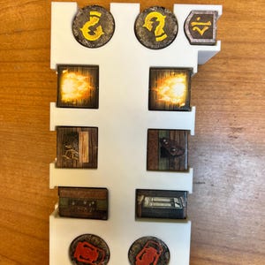 May include: A white plastic game piece organizer holding various game tokens. The tokens feature illustrations of flames, chests, and question marks. The tokens are brown, orange, and yellow. The organizer is rectangular with slots for the tokens.