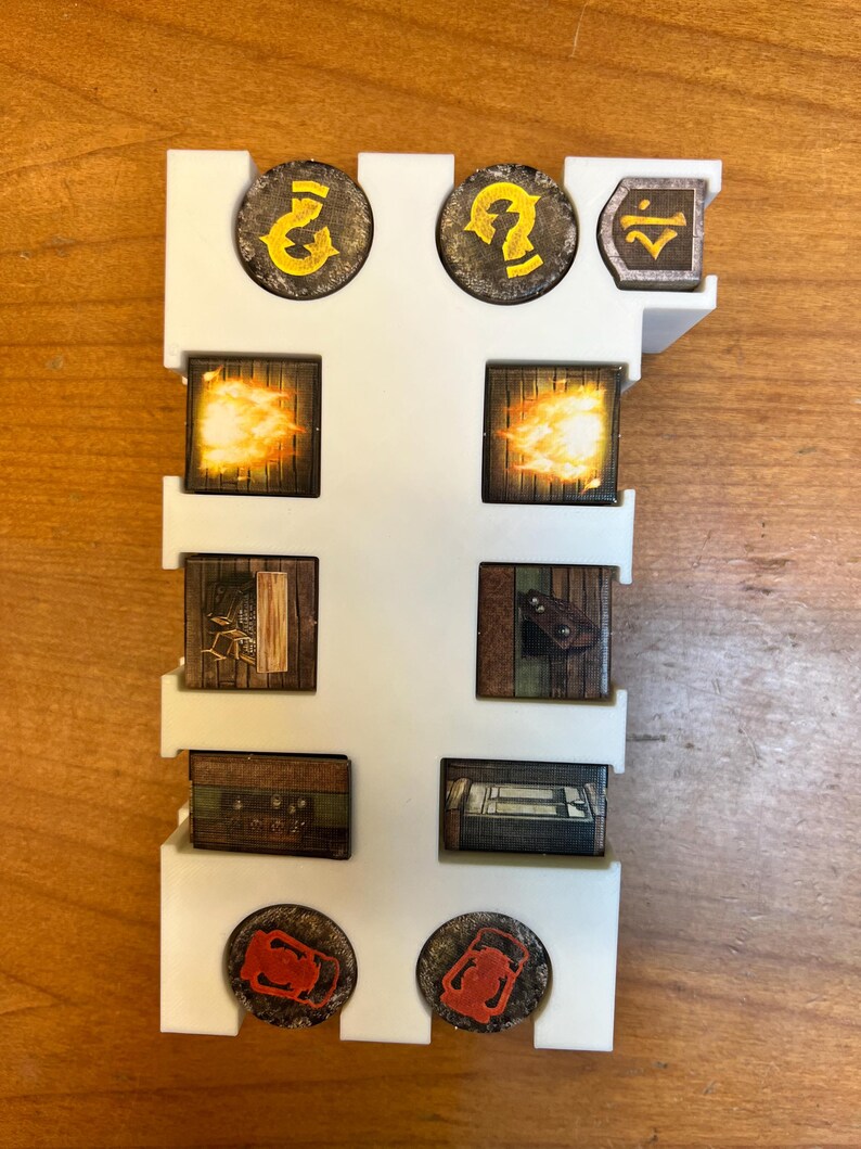 May include: A white plastic game piece organizer holding various game tokens. The tokens feature illustrations of flames, chests, and question marks. The tokens are brown, orange, and yellow. The organizer is rectangular with slots for the tokens.