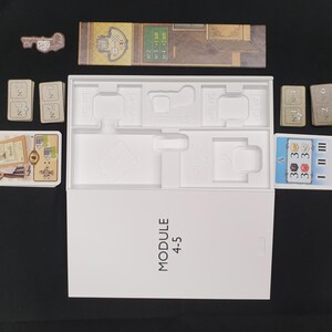 May include: A board game set with various components, including cards, game boards, and a key-shaped token. The white box is labeled "MODULE 4-5". The game pieces are arranged on a black surface.