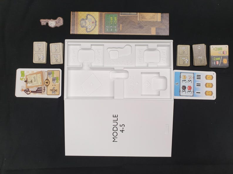 May include: A board game set with various components, including cards, game boards, and a key-shaped token. The white box is labeled "MODULE 4-5". The game pieces are arranged on a black surface.