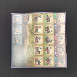 May include: A board game with illustrated tiles depicting ships, buildings, and landscapes. The tiles are arranged in a rectangular box with a purple border. The game's theme appears to be historical or maritime, with a variety of detailed illustrations.
