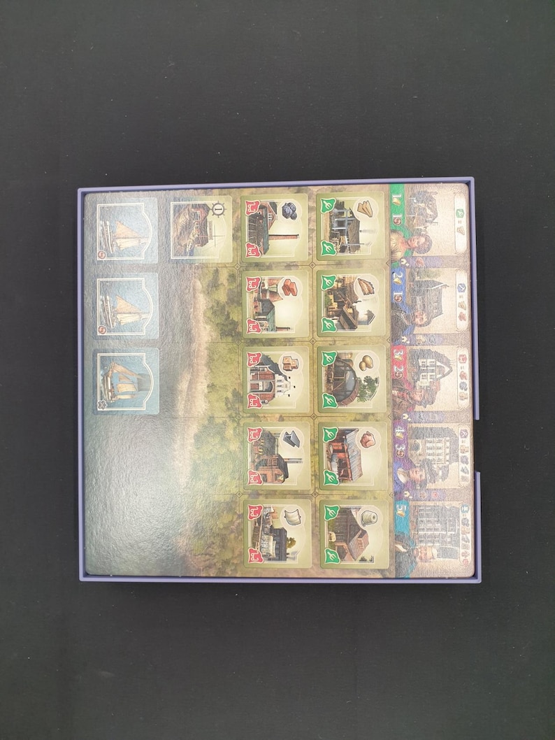 May include: A board game with illustrated tiles depicting ships, buildings, and landscapes. The tiles are arranged in a rectangular box with a purple border. The game's theme appears to be historical or maritime, with a variety of detailed illustrations.