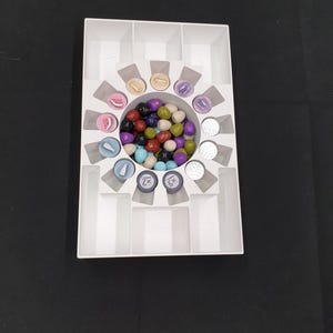 May include: A white plastic game organiser with various game pieces. The organiser has multiple compartments, including a central circular area filled with colourful game tokens. Other pieces include round tokens with symbols and text.