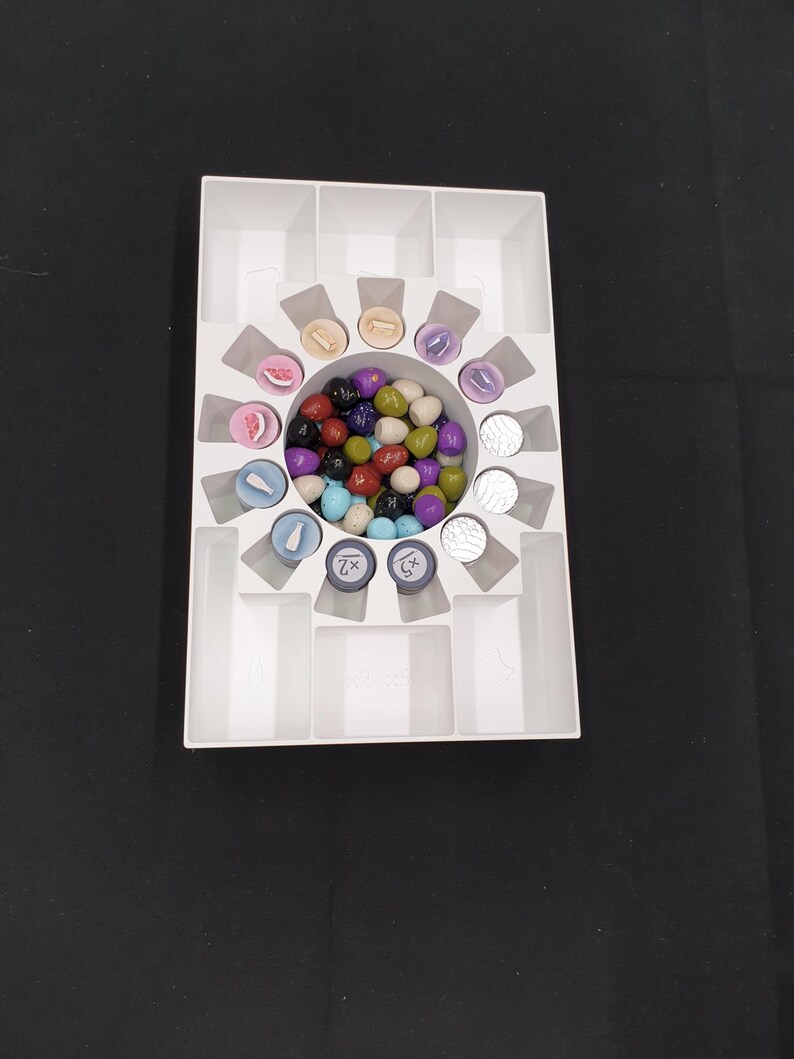 May include: A white plastic game organiser with various game pieces. The organiser has multiple compartments, including a central circular area filled with colourful game tokens. Other pieces include round tokens with symbols and text.