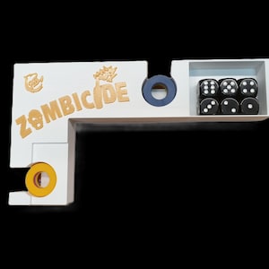 May include: A white wooden game organizer with the word "ZOMBICIDE" in gold lettering. The organizer has compartments for dice and other game components. Six black dice with white dots are visible.