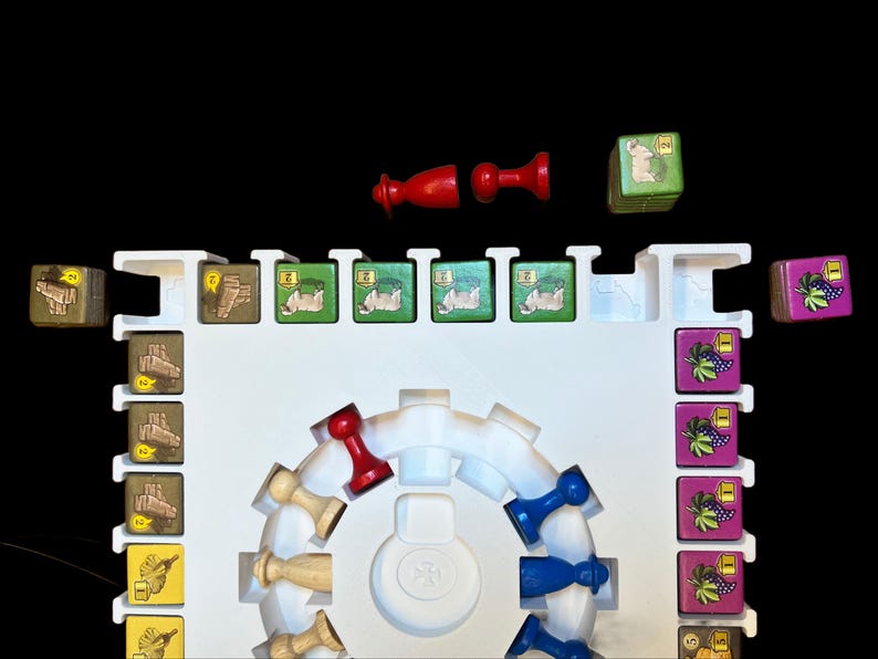May include: A white game board with a circular track in the center. There are red, blue, and wooden game pieces on the track. There are also small square game tiles with images of sheep, grapes, and other items. The tiles are arranged in rows on the board.