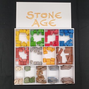 Puede incluir: Una caja organizadora blanca con las palabras "STONE AGE" en letras doradas. La caja est&aacute; dividida en compartimentos llenos de piezas de juego coloridas, incluyendo formas amarillas, verdes, rojas y azules, dados y fichas. Las piezas son de madera y pl&aacute;stico.