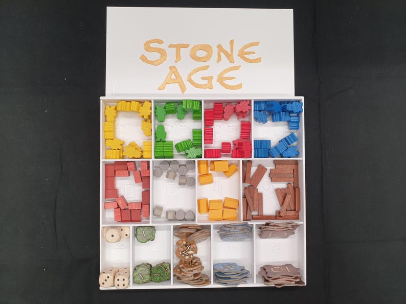 Puede incluir: Una caja organizadora blanca con las palabras "STONE AGE" en letras doradas. La caja est&aacute; dividida en compartimentos llenos de piezas de juego coloridas, incluyendo formas amarillas, verdes, rojas y azules, dados y fichas. Las piezas son de madera y pl&aacute;stico.