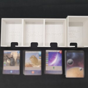 May include: A white plastic card organiser with four compartments, each labelled with text. The organiser holds several stacks of cards with colourful images. The text on the organiser reads: "ARRAKIS LIAISON", "THE SPICE MUST FLOW", "FOLDSPACE", and "IMPERIUM DECK CARDS".