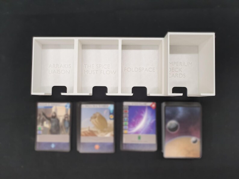 May include: A white plastic card organiser with four compartments, each labelled with text. The organiser holds several stacks of cards with colourful images. The text on the organiser reads: "ARRAKIS LIAISON", "THE SPICE MUST FLOW", "FOLDSPACE", and "IMPERIUM DECK CARDS".