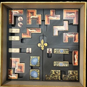 May include: A black foam insert with compartments for storing game pieces. The insert features a fleur-de-lis design in the center and is designed to fit inside a game box.